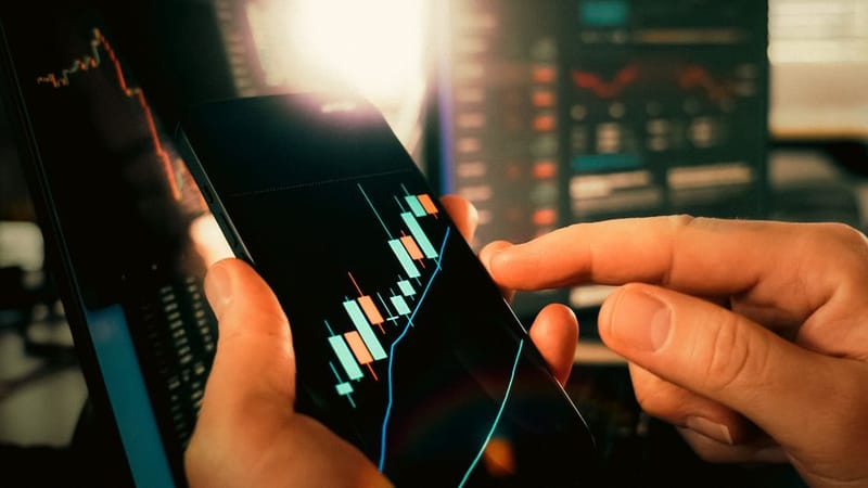 Hand holding a phone showing a stock trading app with candlestick charts